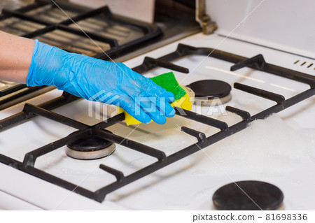 Hand of a woman in a household rubber glove sponges the grate of a gas stove and burners on kitchen, close up 81698336