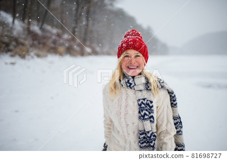 Front view portrait of happy senior woman with hat outdoors standing in snowy nature, looking at camera. 81698727