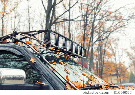 Close-up detail view of custom made roof rack bar with extra headlight mounted on roof of heavy duty pick up suv car against foggy autumn forest. Fallen leaves vehicle windshield. Fall weather drive Close-up detail view of custom made roof rack bar with extra headlight mounted on roof of heavy duty pick up suv car against foggy autumn forest. Fallen leaves vehicle windshield. Fall weather drive 81699093