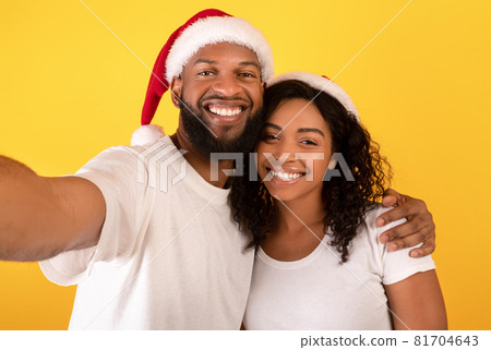 Loving african american couple in Santa hats smiling and taking selfie on yellow studio background, free space Loving african american couple in Santa hats smiling and taking selfie on yellow studio background, free space 81704643