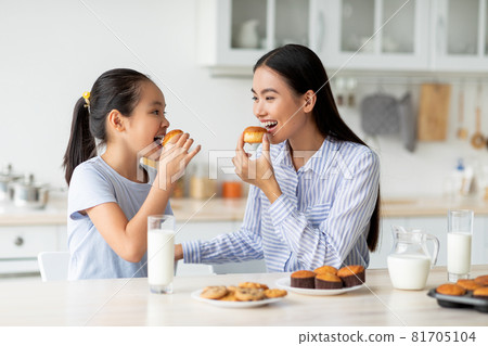 Dessert time for sweet tooth. Happy asian girl and young woman eating cookies, sitting at dinner table in kitchen 81705104