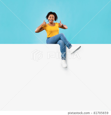 Excited young black lady sitting on blank white paper banner with mockup for ad, showing thumb up on blue background 81705659