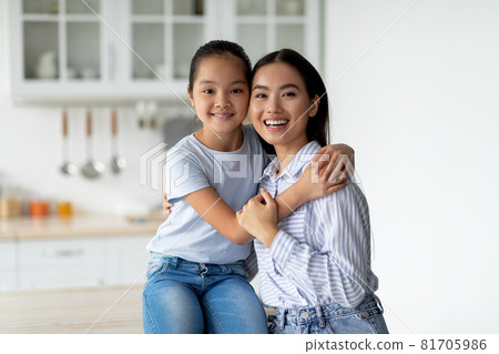 Portrait of young asian mother and cute little daughter posing in kitchen interior, pretty girl hugging her mom 81705986