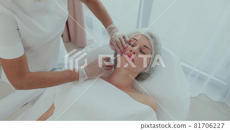 Top view. Attractive woman gets anti-aging face injections. An experienced young cosmetologist fills female wrinkles with hyaluronic acid from a syringe. Top view. Attractive woman gets anti-aging face injections. An experienced young cosmetologist fills female wrinkles with hyaluronic acid from a syringe. 81706227