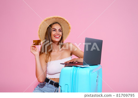 girl with laptop tickets, credit card and passport are going to travel. Sitting near suitcase in shorts white top and straw hat. Pink background. looking at laptop going to book something girl with laptop tickets, credit card and passport are going to travel. Sitting near suitcase in shorts white top and straw hat. Pink background. looking at laptop going to book something 81709795