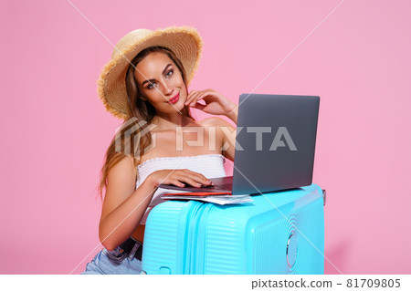 Smiling girl with laptop tickets money and passport are going to travel. Portrait. Sitting near suitcase in shorts white top and straw hat. Pink background. looks at the camera  81709805