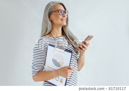 Mature Asian woman with loose hair holds diagrams and mobile phone on light grey background Mature Asian woman with loose hair holds diagrams and mobile phone on light grey background 81710117