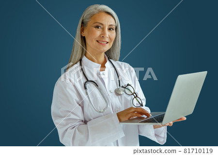 Smiling mature woman doctor in white coat works on laptop standing on blue background Smiling mature woman doctor in white coat works on laptop standing on blue background 81710119
