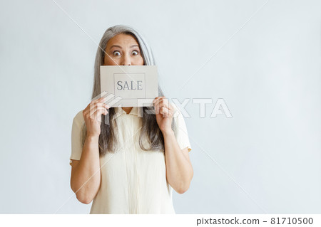 Shocked silver haired Asian lady holds card with word Sale on light grey background Shocked silver haired Asian lady holds card with word Sale on light grey background 81710500