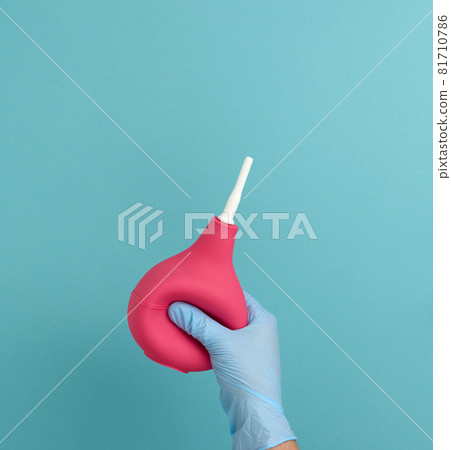 a hand in a blue medical glove holds a pink rubber enema on a blue background 81710786