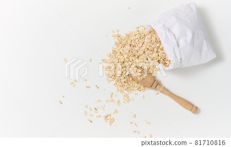 raw oatmeal in a white paper bag and a wooden spoon on a white table, breakfast porridge 81710816