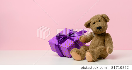 cute brown teddy bear holding a box wrapped in paper and red silk ribbon on white table. Prize and congratulations, 81710822