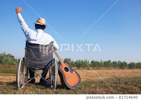 Young disabled man and field background. He is raising guitar and looking into sky.He is wearing a hat and sitting on wheelchair.despair,lonely,hope.Photo concept depression and Patient. Young disabled man and field background. He is raising guitar and looking into sky.He is wearing a hat and sitting on wheelchair.despair,lonely,hope.Photo concept depression and Patient. 81714640