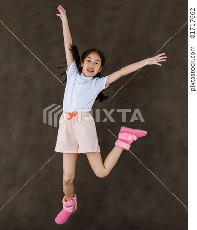 Portrait studio shot of little cute playful happy Asian girl wears white shirt and pink shorts and boots jumping high showing peace sign with two fingers overhead in front of brown wall background Portrait studio shot of little cute playful happy Asian girl wears white shirt and pink shorts and boots jumping high showing peace sign with two fingers overhead in front of brown wall background 81717662