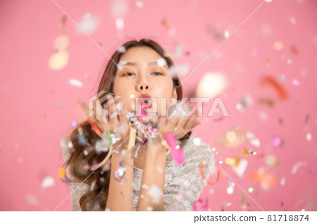 Portrait of a cheerful beautiful Asian womanl wearing dress standing standing under confetti rain and celebrating isolated over pink background.She blowing confetti from her hands . Portrait of a cheerful beautiful Asian womanl wearing dress standing standing under confetti rain and celebrating isolated over pink background.She blowing confetti from her hands . 81718874