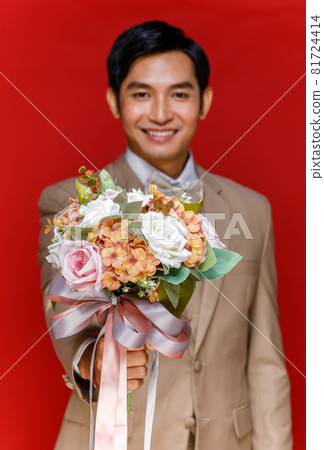 Young attractive Asian man soon to be groom wearing white shirt and beige vest and suit holding bouquet of flower against red background. Concept for pre wedding photography 81724414