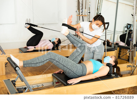 Female pilates trainer in protective mask helping pilates woman during training Female pilates trainer in protective mask helping pilates woman during training 81726889