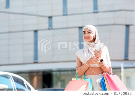 Smiling emotional millennial arab lady in hijab with a lot of packages and phone looks at empty space 81730983