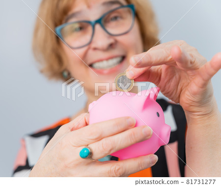 An elderly woman with glasses puts a coin in a piggy bank on a white background An elderly woman with glasses puts a coin in a piggy bank on a white background 81731277
