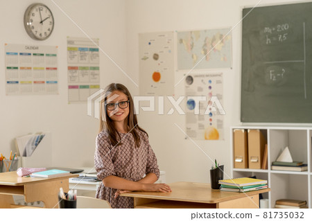 Adorable schoolgirl in dress and eyeglasses standing by desk Adorable schoolgirl in dress and eyeglasses standing by desk 81735032