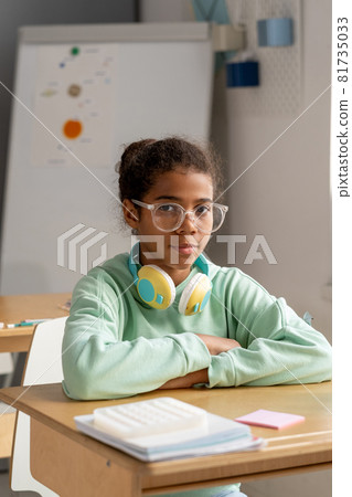 Cute serious schoolgirl in eyeglasses sitting by her desk Cute serious schoolgirl in eyeglasses sitting by her desk 81735033