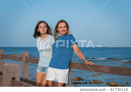 Mother and daughter standing on wooden sidewalk by the sea at sunset Mother and daughter standing on wooden sidewalk by the sea at sunset 81736118