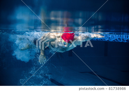 Underwater view of professional male swimmer in red cap and goggles in motion and action during training at pool, indoors. Healthy lifestyle, power, energy, sports movement concept 81738986