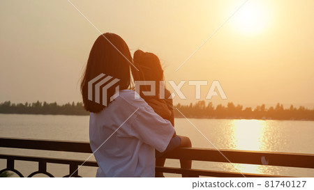 Mother holds little girl on railing on waterfront at sunset Mother holds little girl on railing on waterfront at sunset 81740127