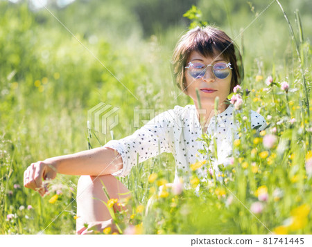 Woman in colorful sunglasses, enjoys sunlight and flower fragrance on grass field. Summer vibes. Relax outdoors. Self-soothing. 81741445