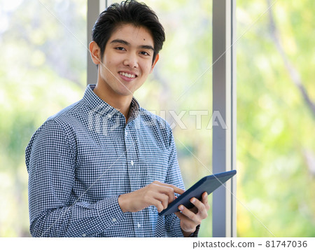 Portrait shot of Asian young happy handsome successful male businessman standing smiling look at camera holding touch screen tablet in hands in front glass windows with blurred green tree background 81747036