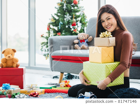 Asian young happy beautiful female sitting on carpet floor surround by colorful presents and teddy bear dolls smiling look at camera hold wrapped gift boxes on lap in front decoration Christmas tree 81747146