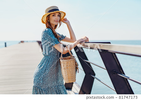 Beautiful redhead woman posing at sunny day. walks along the pier near the sea. Girl wearing fashionable blue dress and straw hat. Holding straw handbag. Looking at the camera Beautiful redhead woman posing at sunny day. walks along the pier near the sea. Girl wearing fashionable blue dress and straw hat. Holding straw handbag. Looking at the camera 81747173