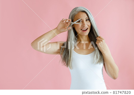 Happy middle aged woman with long hoary hair looks through open scissors in studio Happy middle aged woman with long hoary hair looks through open scissors in studio 81747820