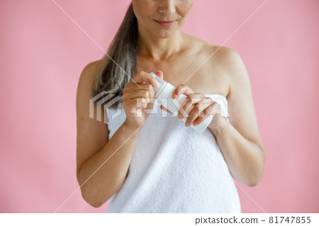Happy middle aged woman with terry towel holds body care product on pink background 81747855