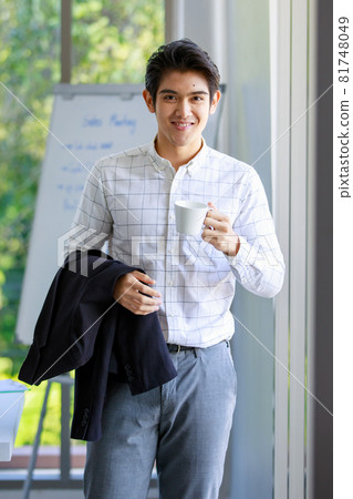 Portrait studio shot of Asian successful happy handsome male sales marketing officer standing smiling look at camera holding coffee cup and suit jacket in hands in meeting room in front white board 81748049