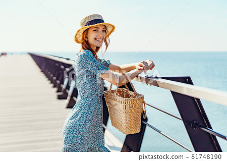 Beautiful redhead woman posing at sunny day. walks along the pier near the sea. Girl wearing fashionable blue dress and straw hat. Holding straw handbag. Looking at the camera. Smiling. Laughing Beautiful redhead woman posing at sunny day. walks along the pier near the sea. Girl wearing fashionable blue dress and straw hat. Holding straw handbag. Looking at the camera. Smiling. Laughing 81748190