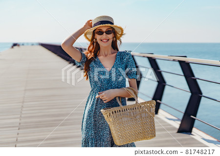 Beautiful redhead woman posing at sunny day. walks along the pier near the sea. Girl wearing fashionable sunglasses blue dress and straw hat. Holding straw handbag. smiling Beautiful redhead woman posing at sunny day. walks along the pier near the sea. Girl wearing fashionable sunglasses blue dress and straw hat. Holding straw handbag. smiling 81748217