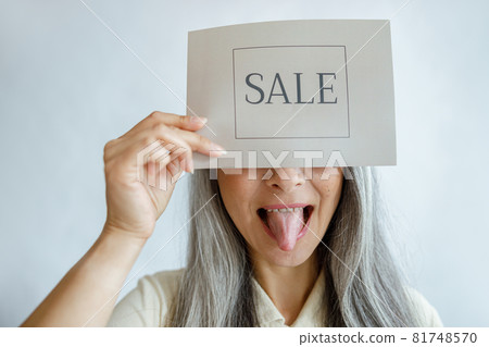 Playful lady with silver hair holds card with word Sale and shows tongue in studio Playful lady with silver hair holds card with word Sale and shows tongue in studio 81748570