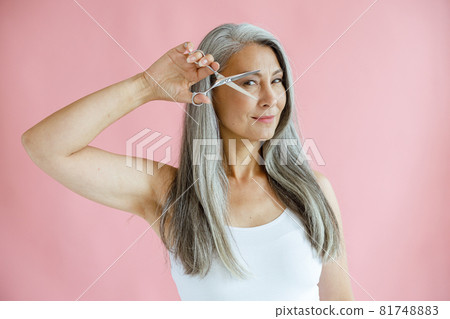 Beautiful Asian lady with grey hair holds open scissors near face on pink background Beautiful Asian lady with grey hair holds open scissors near face on pink background 81748883