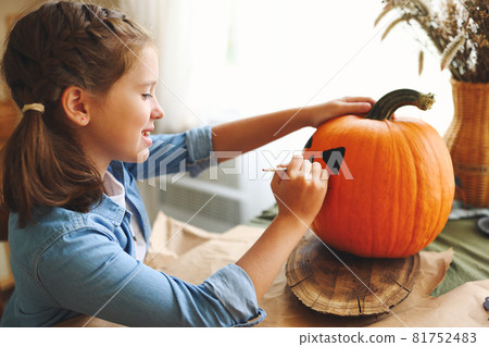 Smiling little kids girl making jack-o-lantern at home, drawing scary face on pumpkin 81752483
