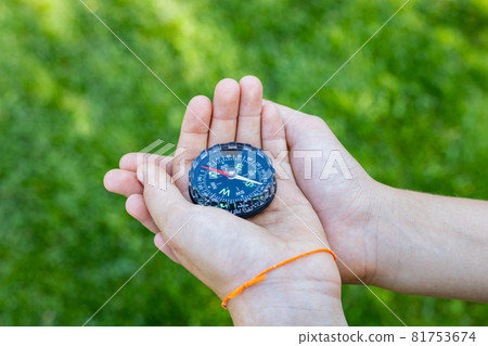 Girl's hands holding the compass. Hands of a teenager girl holding a liquid compass. Red compass needle points north. Green grass background. Copy space. Orienteering on the ground. Girl's hands holding the compass. Hands of a teenager girl holding a liquid compass. Red compass needle points north. Green grass background. Copy space. Orienteering on the ground. 81753674