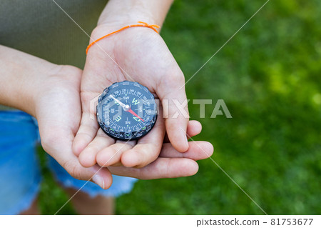 Girls hands holding the compass. Hands of a teenager girl holding a liquid compass. Red compass needle points north. Green grass background. Copy space. Orienteering on the ground. Direction of motion Girls hands holding the compass. Hands of a teenager girl holding a liquid compass. Red compass needle points north. Green grass background. Copy space. Orienteering on the ground. Direction of motion 81753677