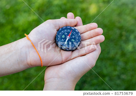 Girl holding the compass. Hands of a teenager girl holding a liquid compass. Green grass background. Copy space. Red compass needle points north. Orienteering on the ground. Selecting the direction Girl holding the compass. Hands of a teenager girl holding a liquid compass. Green grass background. Copy space. Red compass needle points north. Orienteering on the ground. Selecting the direction 81753679