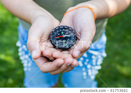 Girl holding the compass. Hands of a teenager holding a liquid compass. Red compass needle points north. Green grass background. Copy space. Orienteering on the ground. Selecting the direction Girl holding the compass. Hands of a teenager holding a liquid compass. Red compass needle points north. Green grass background. Copy space. Orienteering on the ground. Selecting the direction 81753681