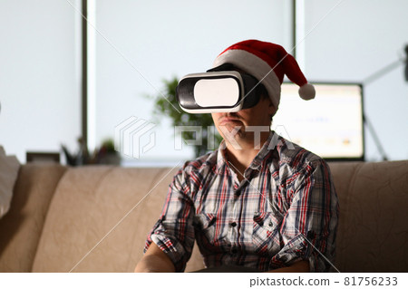 Man in virtual glasses sits on sofa wearing santa claus hat Man in virtual glasses sits on sofa wearing santa claus hat 81756233