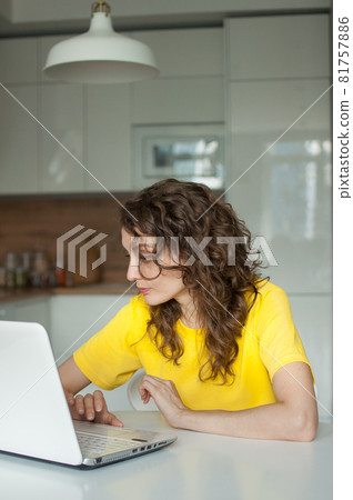 Young woman with curly hair and yellow shirt is working from home using her laptop at the kitchen table in her apartment, remote work, freelance Young woman with curly hair and yellow shirt is working from home using her laptop at the kitchen table in her apartment, remote work, freelance 81757886
