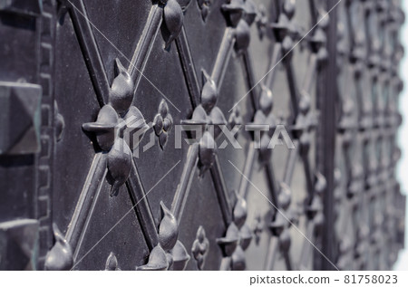 Massive antique black metal door. Surface with repeating ornamentation. Wrought rhombus and geometric pattern. Selective focus. Side view. Outside. Massive antique black metal door. Surface with repeating ornamentation. Wrought rhombus and geometric pattern. Selective focus. Side view. Outside. 81758023