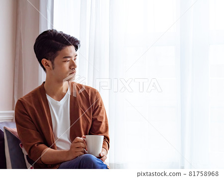 英俊的亞裔年輕人在家裡的窗簾旁邊喝飲料 81758968