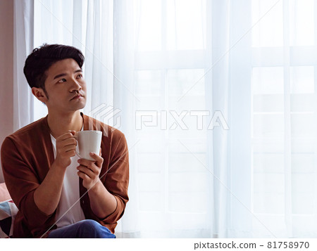 英俊的亞裔年輕人在家裡的窗簾旁邊喝飲料 81758970