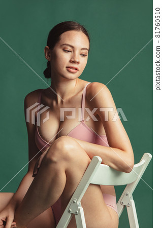 Cropped portrait of young caucasian woman in pink inner wear sitting on chair and looking down isolated over green studio background 81760510